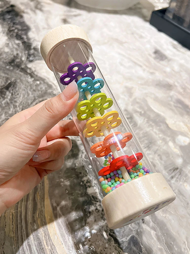 A hand holds a wooden rain sound tube musical instrument with colorful plastic flowers inside.