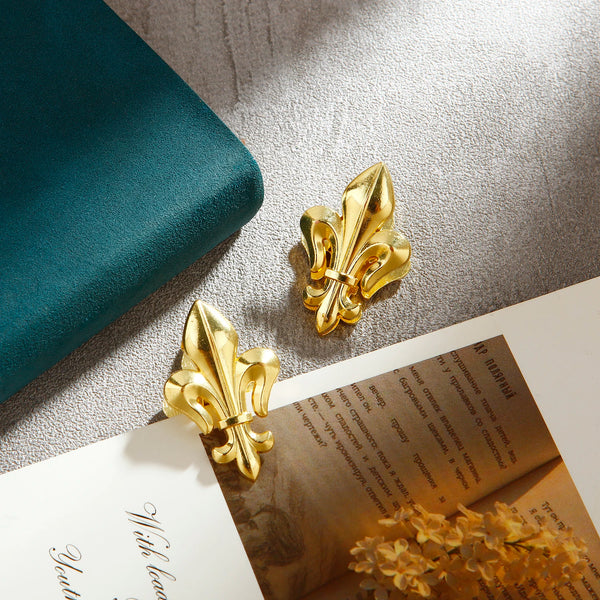 A pair of gold-colored, iris-shaped metal binder clips resting on a light surface next to a teal object.