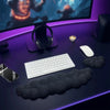 A black ergonomic memory foam wrist rest pad is placed on a purple desk next to a keyboard, mouse, and headphones.