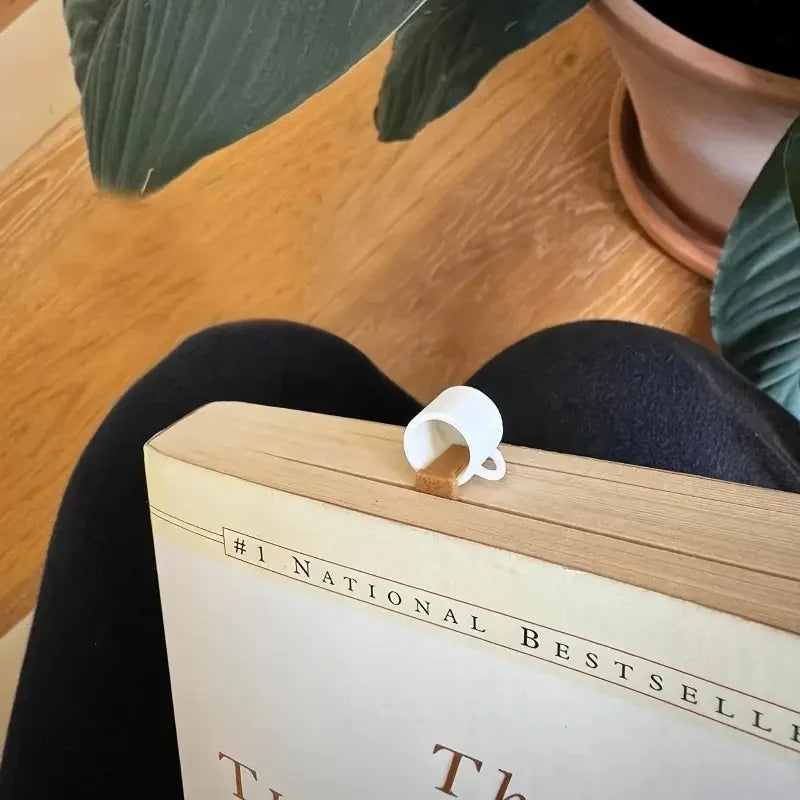 A book with a creative coffee cup bookmark resting on the spine, next to large green leaves.