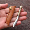 A compact fixed blade knife with a silver blade and brass handle is shown in a brown leather sheath being held in a hand.