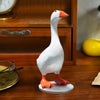 A white resin duck figurine with orange feet standing on a round base.