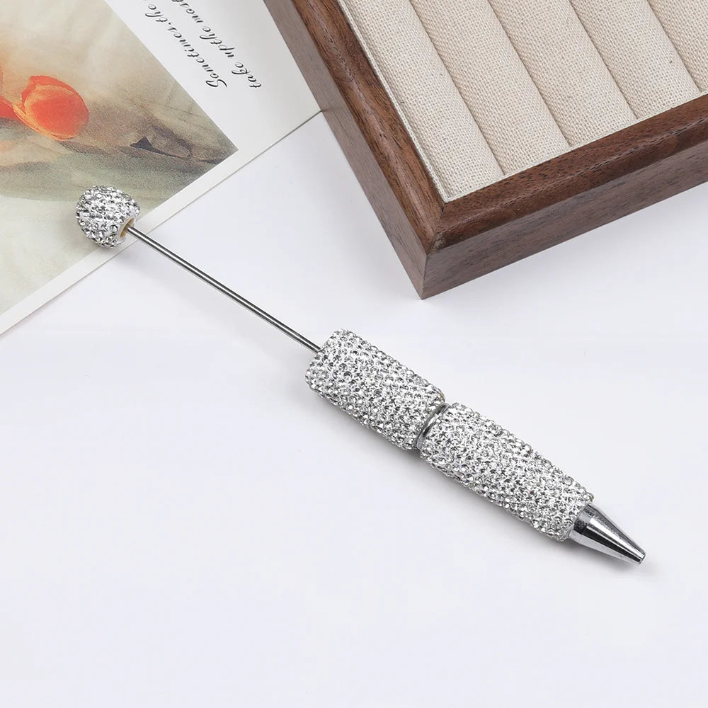 A silver rhinestone beaded pen accessory with a decorative head is displayed on a white surface next to a wooden box and a bo