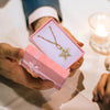 Hands present a pink jewelry gift box with a gold necklace inside, set against a white background.