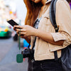 A person holds a smartphone while wearing a tan jacket and a black camera, with a silicone earphone case hanging from the pho