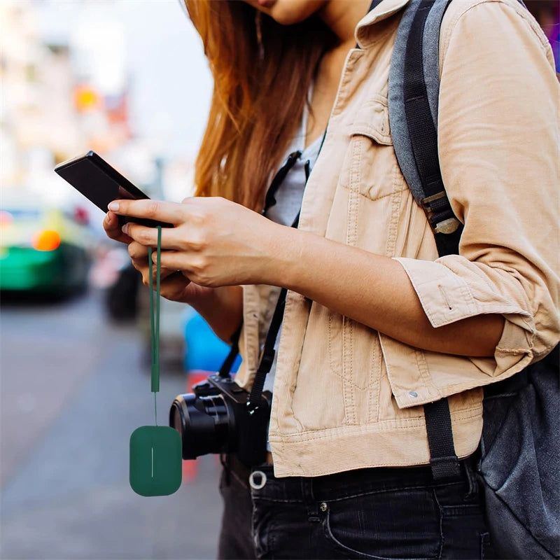 A person holds a smartphone while wearing a tan jacket and a black camera, with a silicone earphone case hanging from the pho