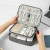 A hand opens a gray travel cable organizer bag showing various white charging cables and electronics storage inside.