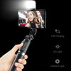 A hand holds a Bluetooth selfie stick with an LED fill light and tripod stand, illuminating a smartphone screen.
