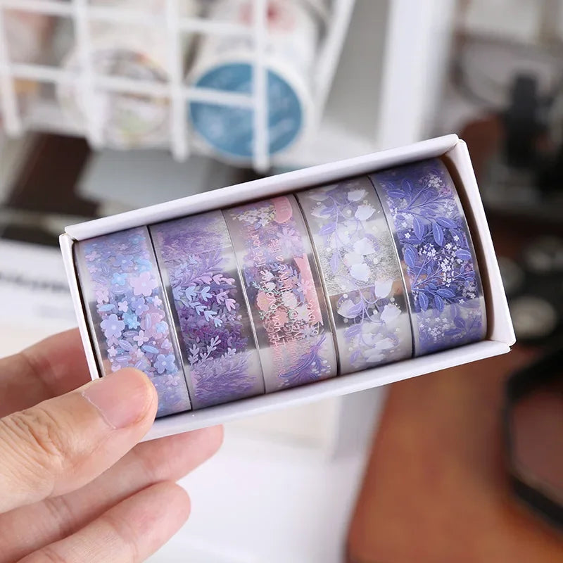 A hand holds a white box containing a set of decorative transparent washi tape rolls with floral patterns.