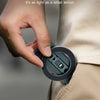 A hand holds a small, dark gray magnetic phone tripod stand with a hook against a light beige fabric.