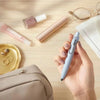 A hand holds a light blue Uni-Ball One Mini Cute Gel Pen on a wooden surface with various cosmetic and stationery items.