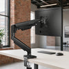 A black premium single monitor desk mount with a gas spring arm is positioned on a light wooden desk.