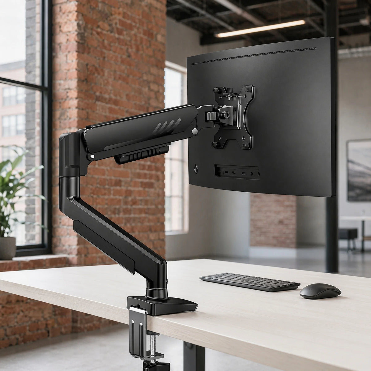 A black premium single monitor desk mount with a gas spring arm is positioned on a light wooden desk.