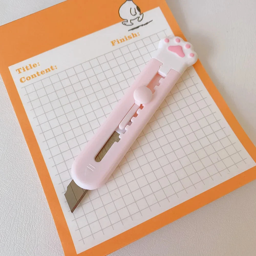 A pink cat paw mini cutter tool is shown resting on a sheet of graph paper with writing for title and content.