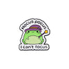 An enamel pin featuring a green frog wearing a purple hat and the text Can't focus.