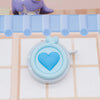 Una cinta métrica compacta y retráctil en forma de corazón, con cuerpo azul claro y un juguete de animal morado de fondo.