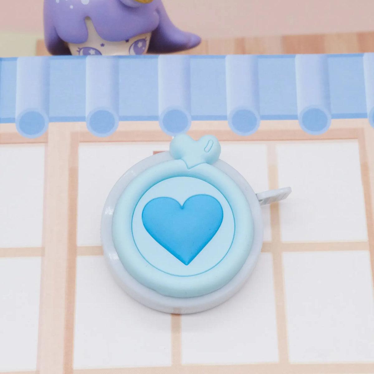 Una cinta métrica compacta y retráctil en forma de corazón, con cuerpo azul claro y un juguete de animal morado de fondo.