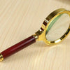 A gold-framed magnifying glass with a dark red handle rests on a light wooden surface.
