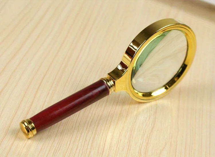 A gold-framed magnifying glass with a dark red handle rests on a light wooden surface.