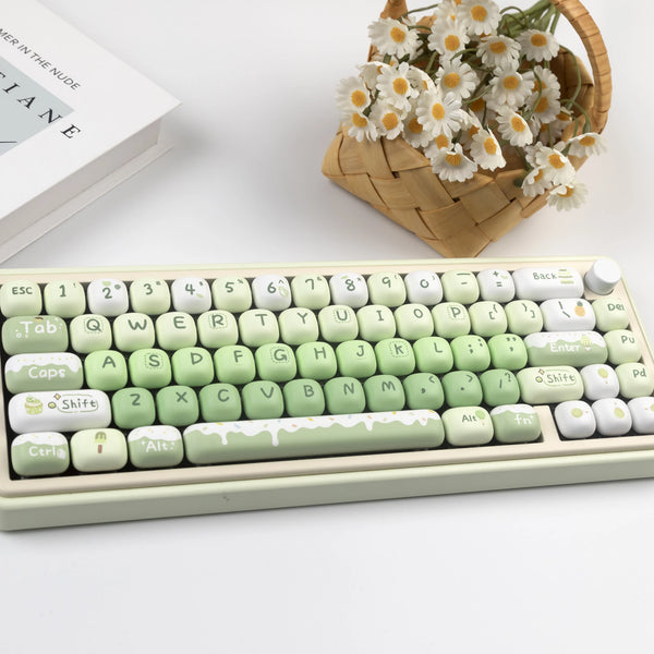 A light green PBT keycap set with custom labels and a wooden basket of white flowers is displayed on a white surface.