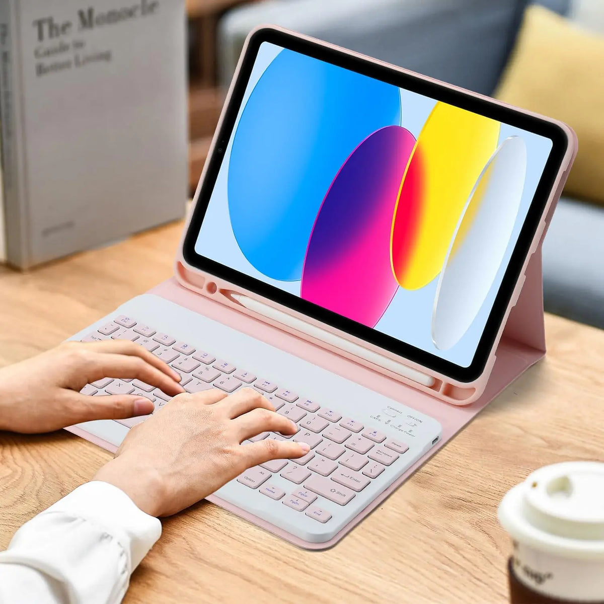 Eine Person tippt auf einem rosa Laptop mit kabellosem Tastatur- und Maus-Set neben einem Buch.