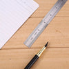 A black pen rests on a wooden surface next to a stainless steel measuring ruler and a sheet of paper.