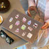 A brown silicone mat features a pack of waterproof cartoon stickers for journaling and decor, held by a person.