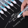 A hand holds a white permanent paint marker pen writing 'Good night happy day' on a black surface with several other pens vis