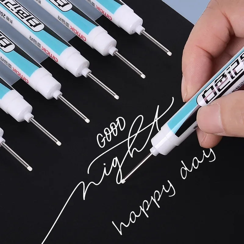 A hand holds a white permanent paint marker pen writing 'Good night happy day' on a black surface with several other pens vis