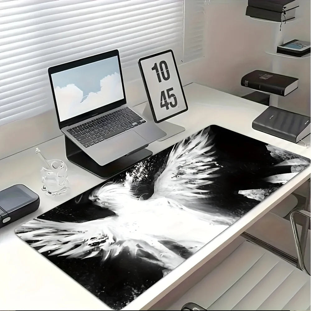 A black and white artistic angel wing desk mat mouse pad is displayed on a white desk next to a laptop.