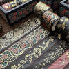 A set of metallic gold washi tape featuring floral and vine patterns on dark paper, displayed next to a wooden box.