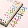 A set of cross-shaped metal paper clips in various colors, laid out next to a page of a notebook.