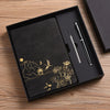 A black box containing a luxury notebook set with gold foil designs and two pens on a wooden surface.