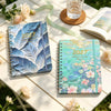 A spiral bound planner with blue and green watercolor designs rests on a wooden table.