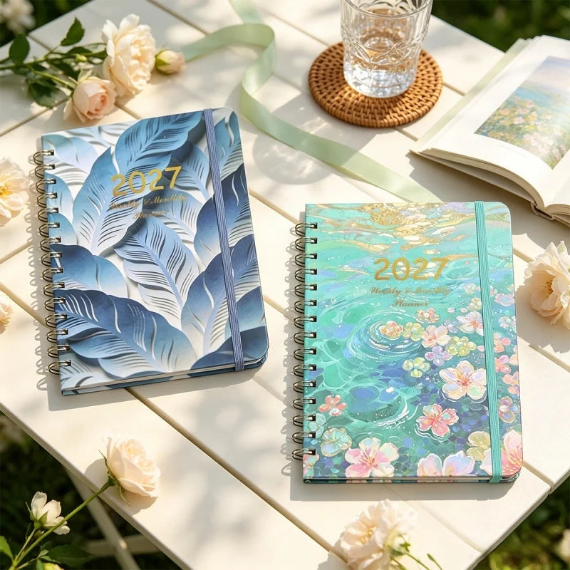 A spiral bound planner with blue and green watercolor designs rests on a wooden table.