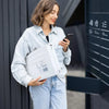 A woman in a denim shirt holds a white self-seal poly mailer box and a smartphone.