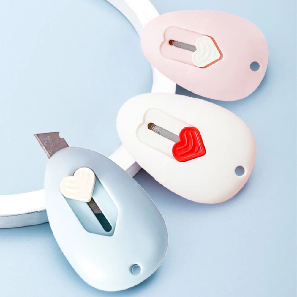 Tres cuchillos de utilidad o herramientas de manualidades rosa y blanco contra un fondo azul claro.