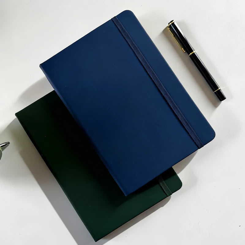 A dark blue A5 hardcover lined notebook with a navy blue ribbon and a black pen resting on a white surface.