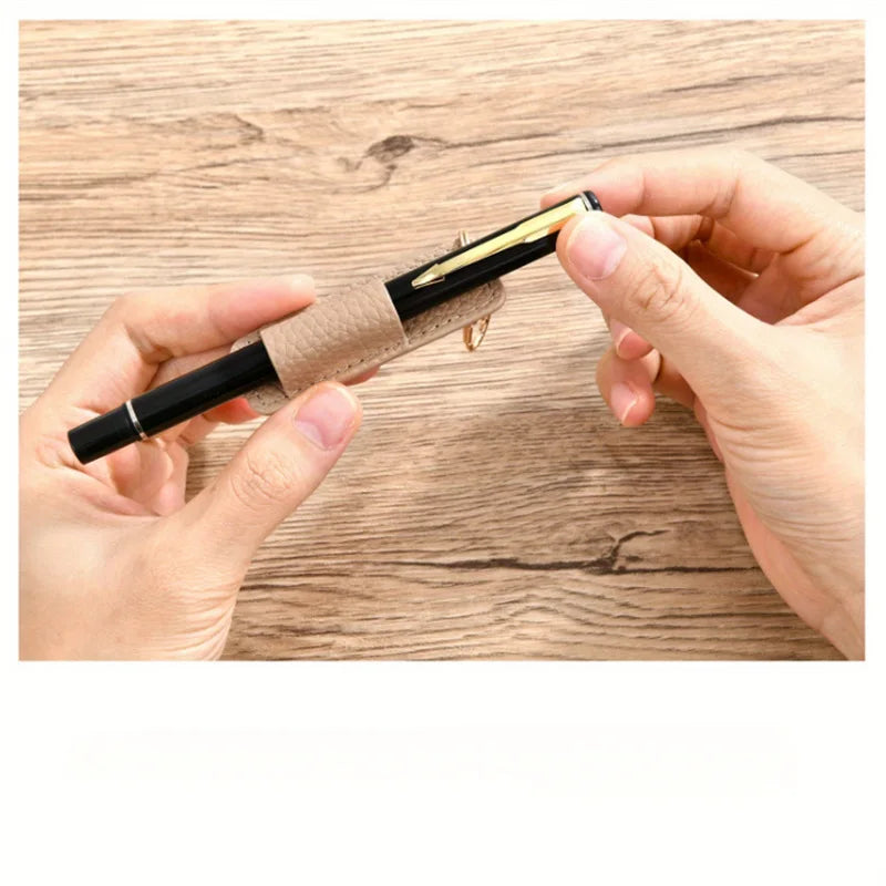 A hand holds a black pen with a leather holder and gold keychain on a wooden surface.