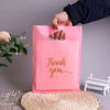 A hand is holding a pink thank you gift bag with a gold script message against a gray background.