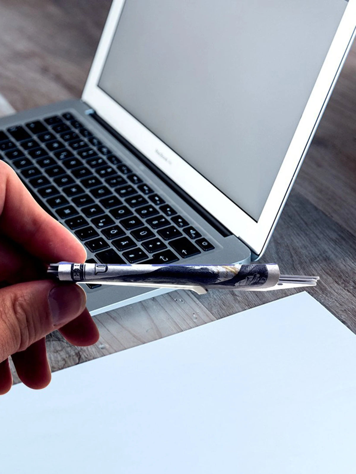 A hand holds an ultra-thin stainless steel money clip wallet next to a silver laptop.