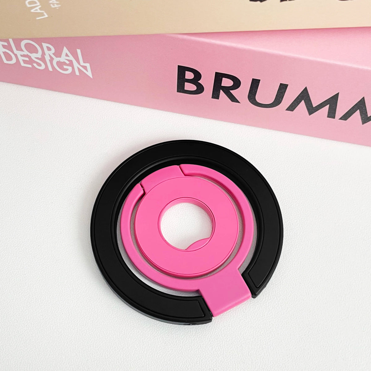 A pink and black magnetic phone grip and stand with a foldable ring resting on a white surface.