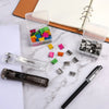 A set of handheld binder clip pushers and stainless steel refill clips are displayed on a white marble surface next to a note