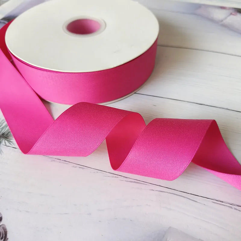 Pink satin ribbon roll with a long strip unwound on a white wooden surface.