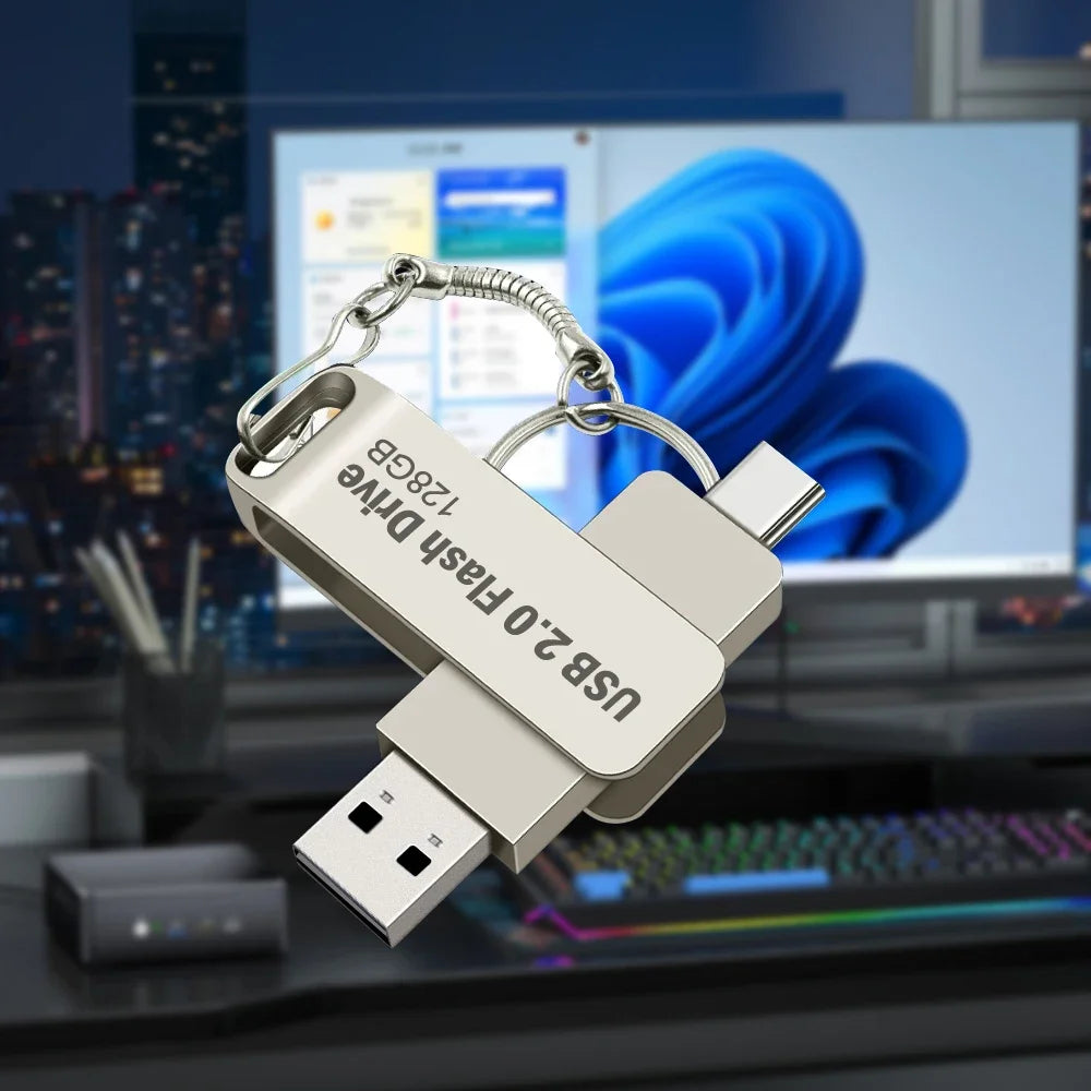 A silver USB 2.0 flash drive is shown with a metal clip, resting on a desk in front of a computer monitor.