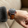 A close-up of a men's watch with a stainless steel case and brown leather band worn on a wrist.