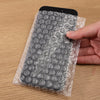 A hand is holding a black smartphone wrapped in clear bubble wrap for padded shipping.