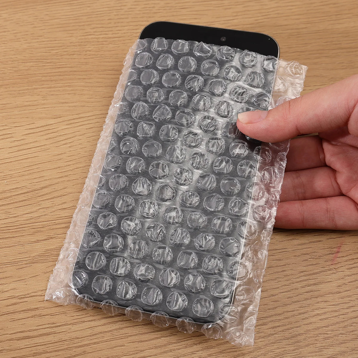 A hand is holding a black smartphone wrapped in clear bubble wrap for padded shipping.
