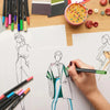 A hand draws a figure with a dual-tip brush marker on white paper next to a set of vibrant markers and color palettes.