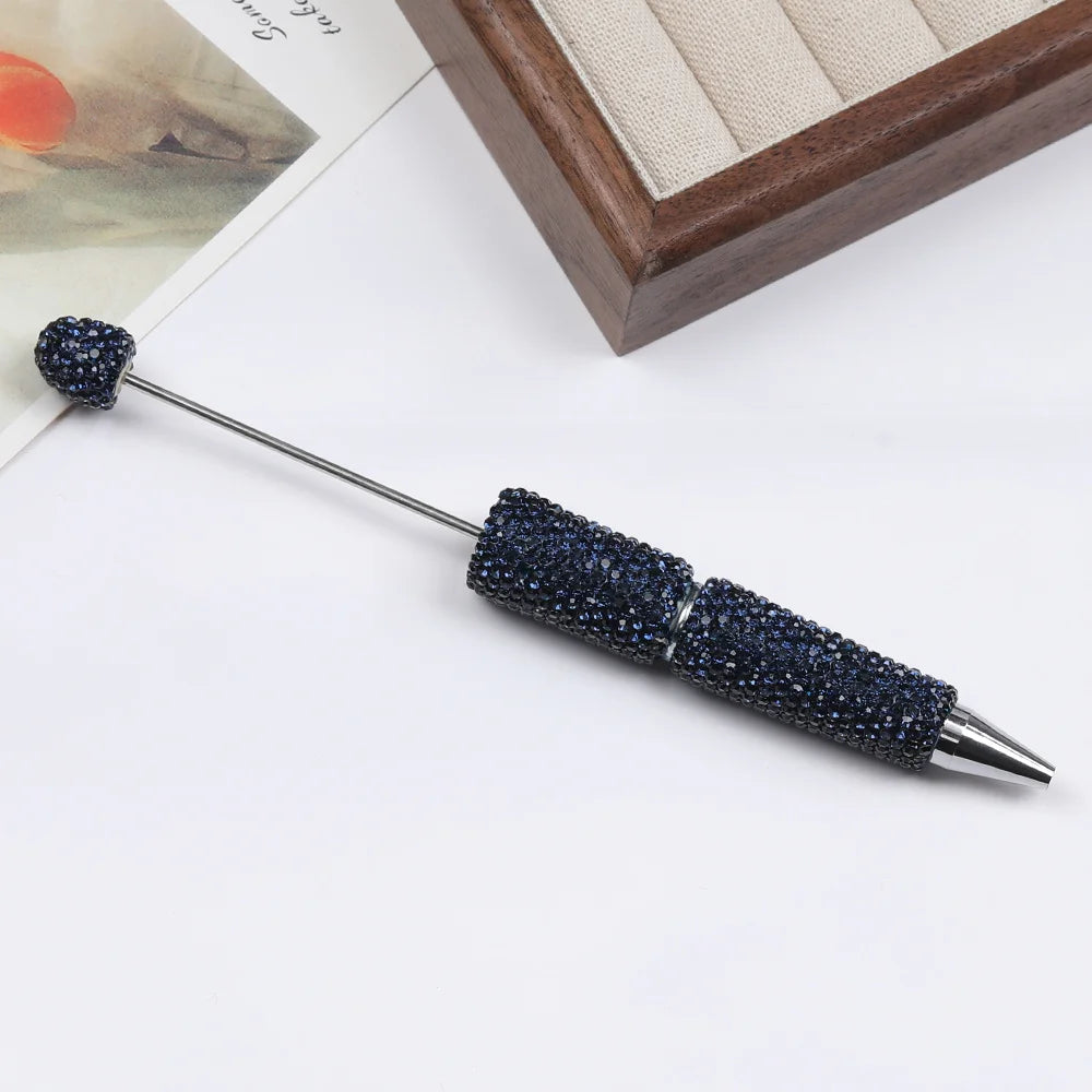 A silver pen accessory with a dark blue rhinestone bead on the tip and a textured, beaded handle.